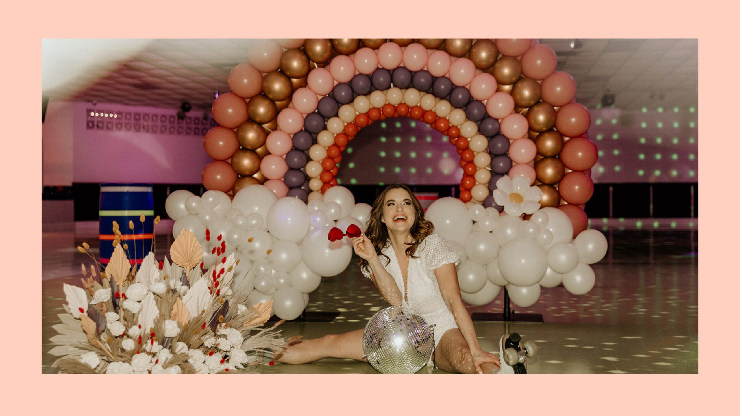 A girl sitting on the floor of a skating rink in front of a large balloon rainbow.