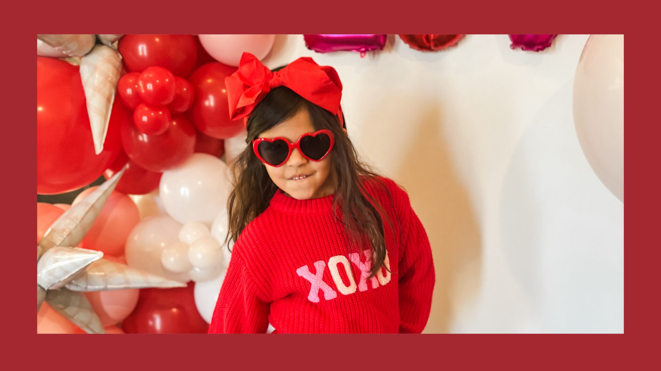 A little girl in a red sweater standing in front of a white backdrop.