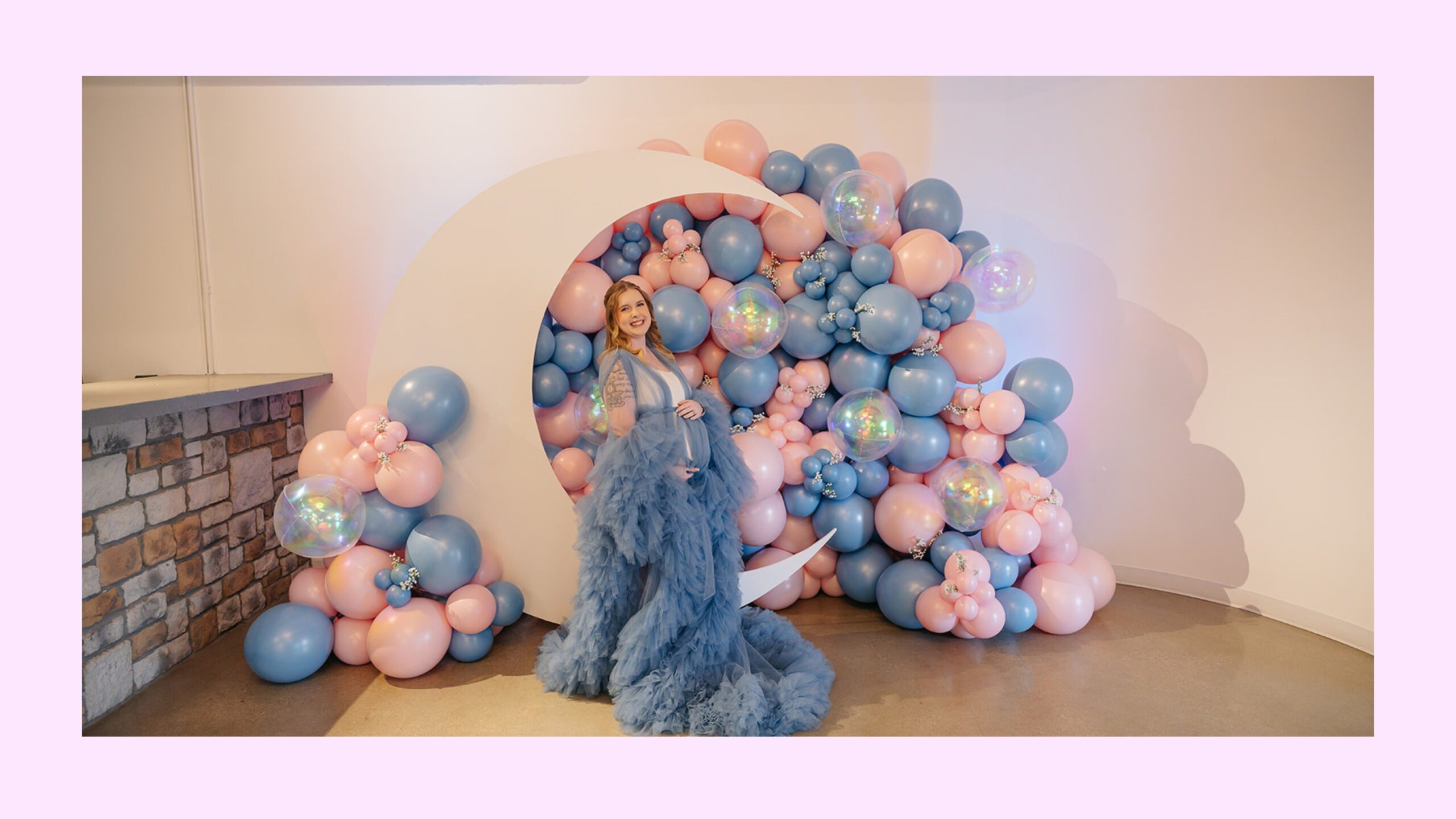 A pregnant woman standing in front of a white moon prop and pink and blue balloons.