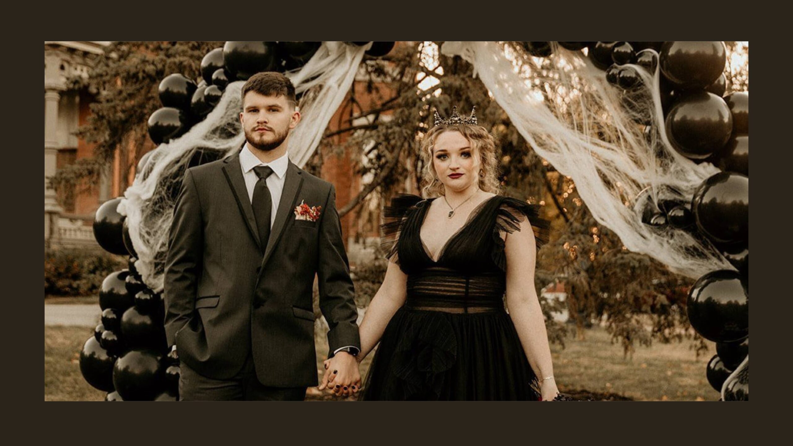 A woman in a black dress and a man in a black suit stand in front of a black balloon arch holding hands.